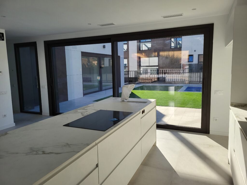 Cocina moderna con ventanales de carpintería de aluminio negro y cristal de gran tamaño. La estancia incluye una isla central de mármol y vistas a un jardín con césped artificial y piscina.